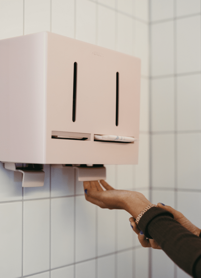 Een persoon gebruikt een tampon/ maandverband dispenser aan de muur in een toilet of badkamer. De dispenser is roze en heeft twee zwarte, verticale lijnen die doen denken aan de ogen van een gezicht, met de papieren handdoeken die eronder uitkomen als een mond. De tegelwand op de achtergrond zorgt voor een schone en hygiënische uitstraling.