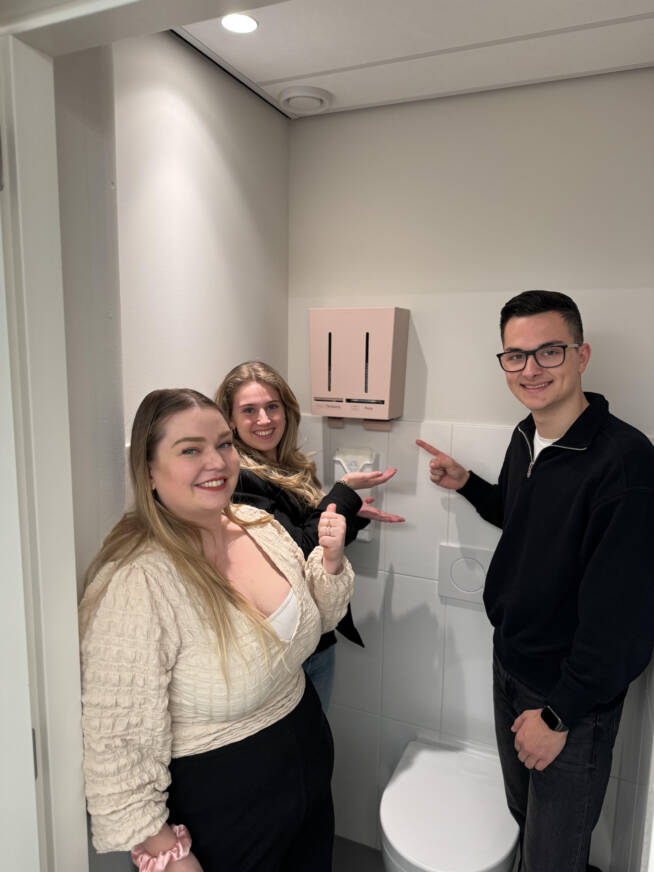 Drie personen, twee vrouwen en een man, staan glimlachend in een toiletruimte en wijzen trots naar een roze dispenser voor menstruatieproducten aan de muur.