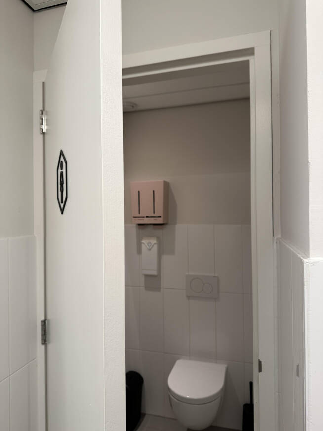 Uitzicht op een open damestoilet met witte tegels en een roze menstruatieproductdispenser aan de muur, naast een witte toiletpot.