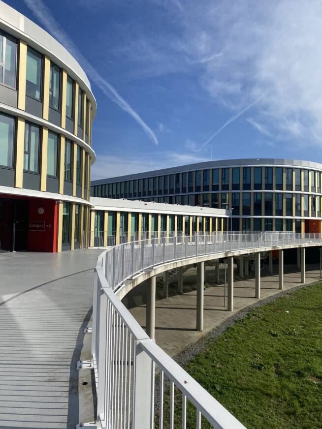 Modern universiteitsgebouw met een gebogen loopbrug op een zonnige dag met blauwe lucht en enkele condensstrepen.