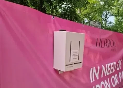 Rosa mensskyddsautomat från Herbox hänger på en mörkrosa reklambanner med Herbox logga i svart