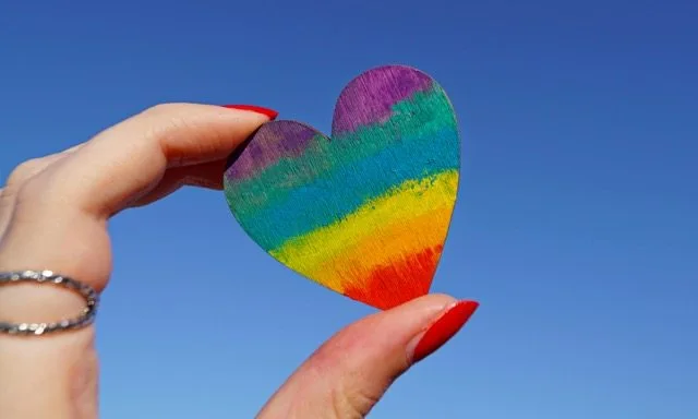 En hand med röda naglar och silverring håller upp ett hjärta målat i prideflaggans färger