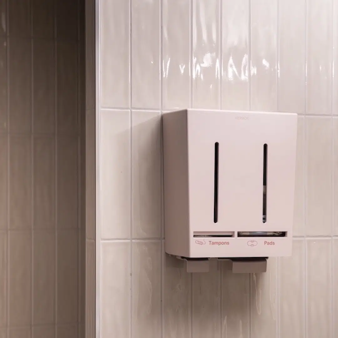 A picture in a restroom of a pink dispenser containing menstrual products.