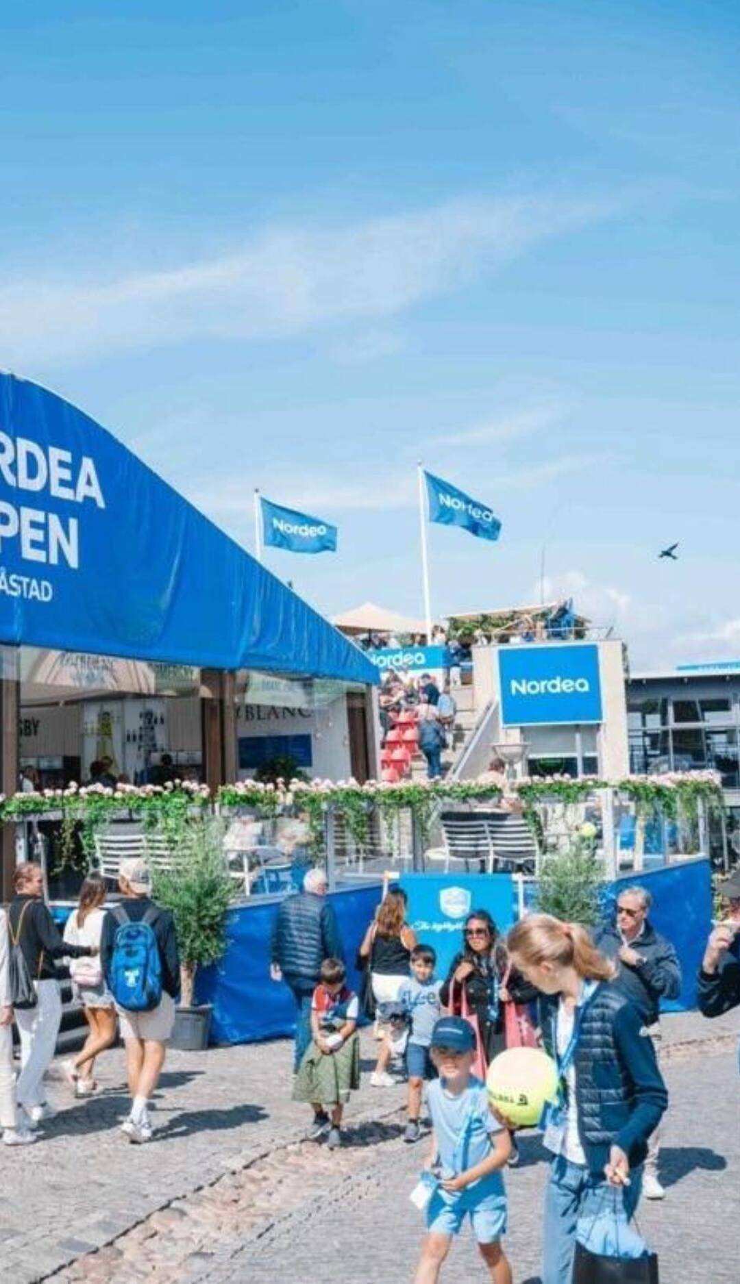 bild på entrén vid tennisveckan i Båstad, flaggstänger med blåa flaggor och Nordeas logga på och människor som går in och ut från arenan