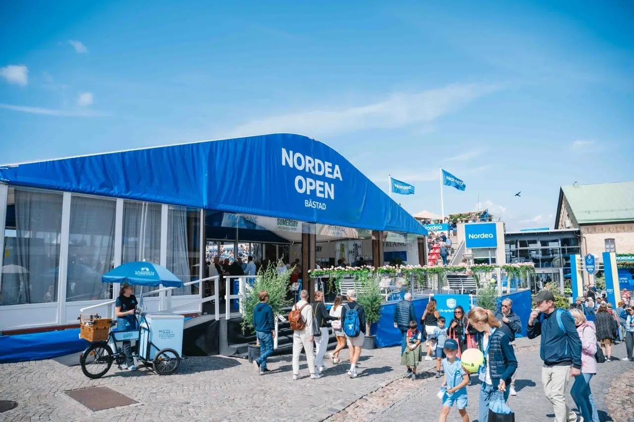 bild på entrén vid tennisveckan i Båstad, flaggstänger med blåa flaggor och Nordeas logga på och människor som går in och ut från arenan