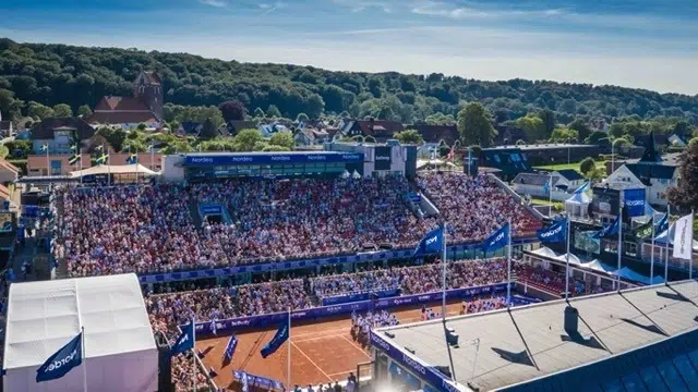 tennisplan i Båstad med en fullsatt publik på läktaren och grön skog i bakgrunden