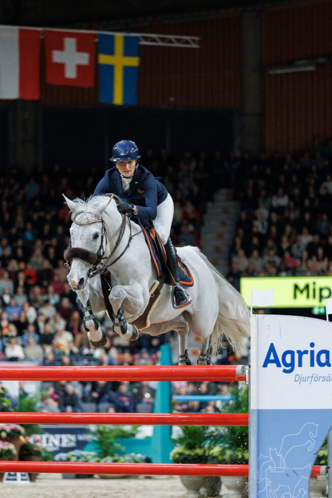 En bild på en tjej/kvinna som rider på en vit häst och hoppar över ett rött hinder i Scandinavium Arena i Göteborg.